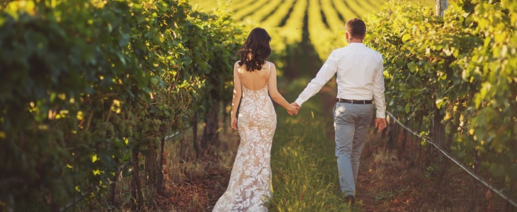 a bride and groom walk away from the camera through a vineyard