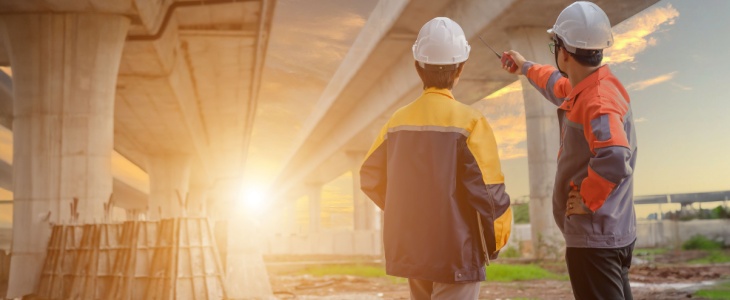 two workers stand together while one gestures across the large construction site